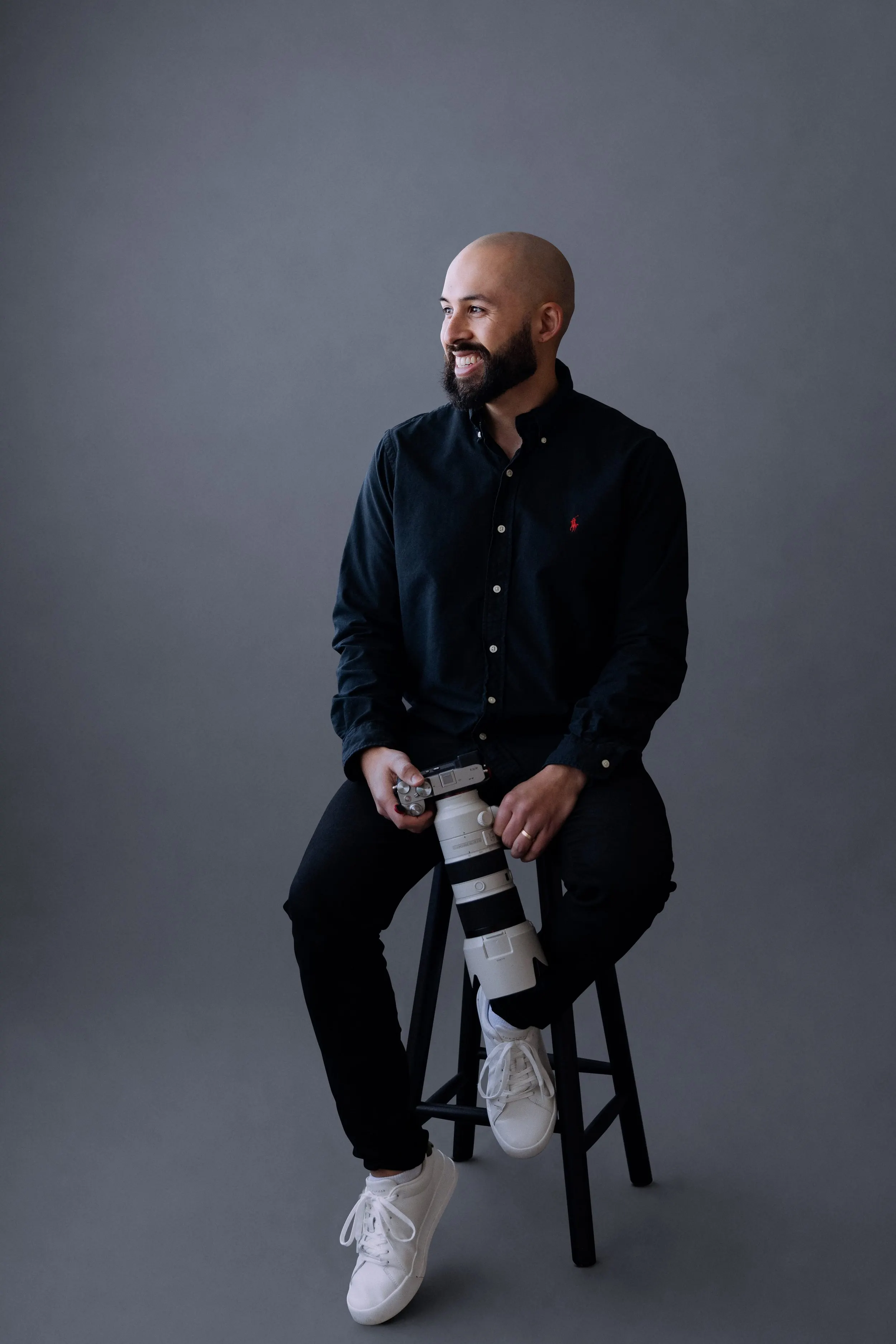 Smiling photographer in a navy shirt seated on a stool with a telephoto lens against a grey backdrop