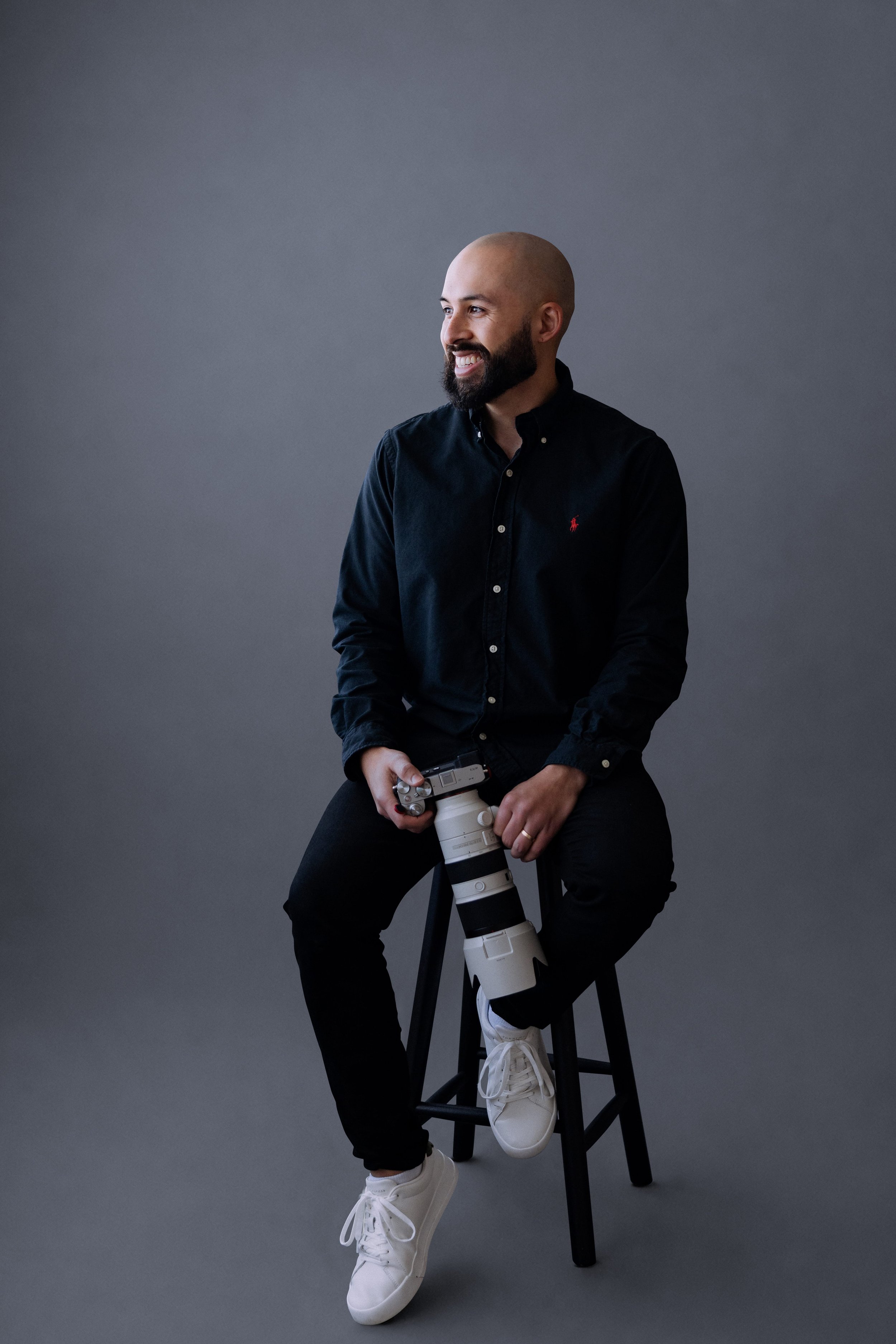 Smiling photographer in a navy shirt seated on a stool with a telephoto lens against a grey backdrop