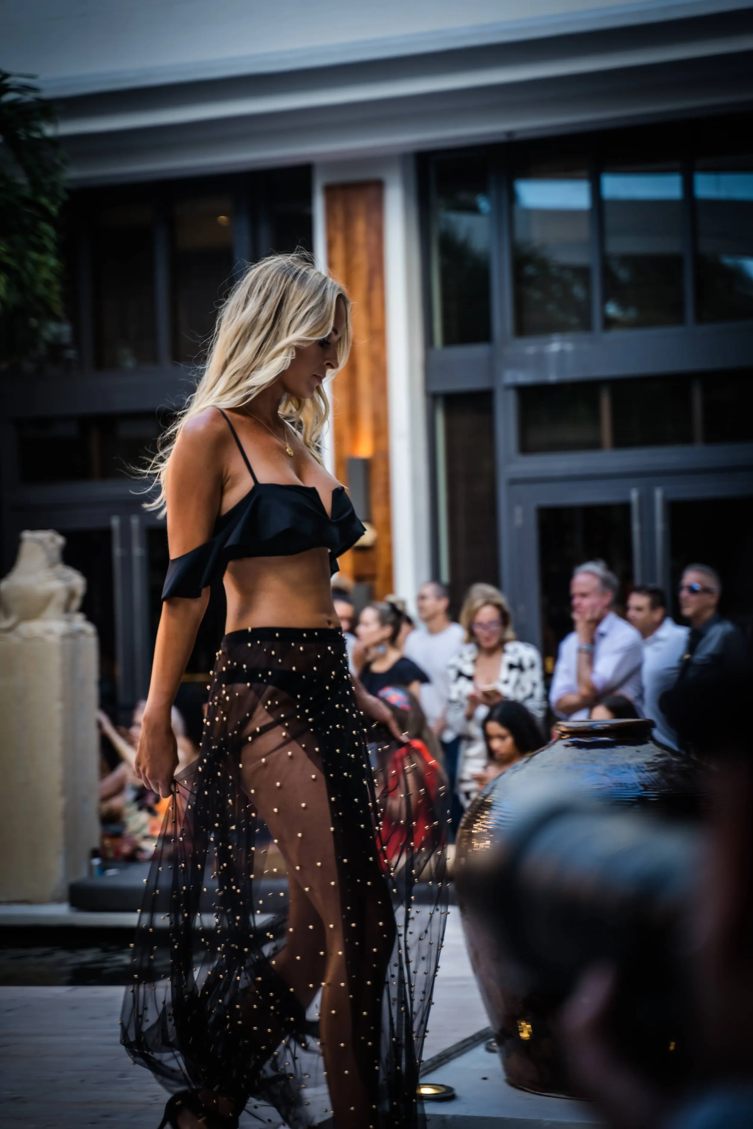 A woman in a black crop top and sheer black skirt with gold embellishments walks on a runway at an outdoor fashion event. Audience members are seated and watching her.