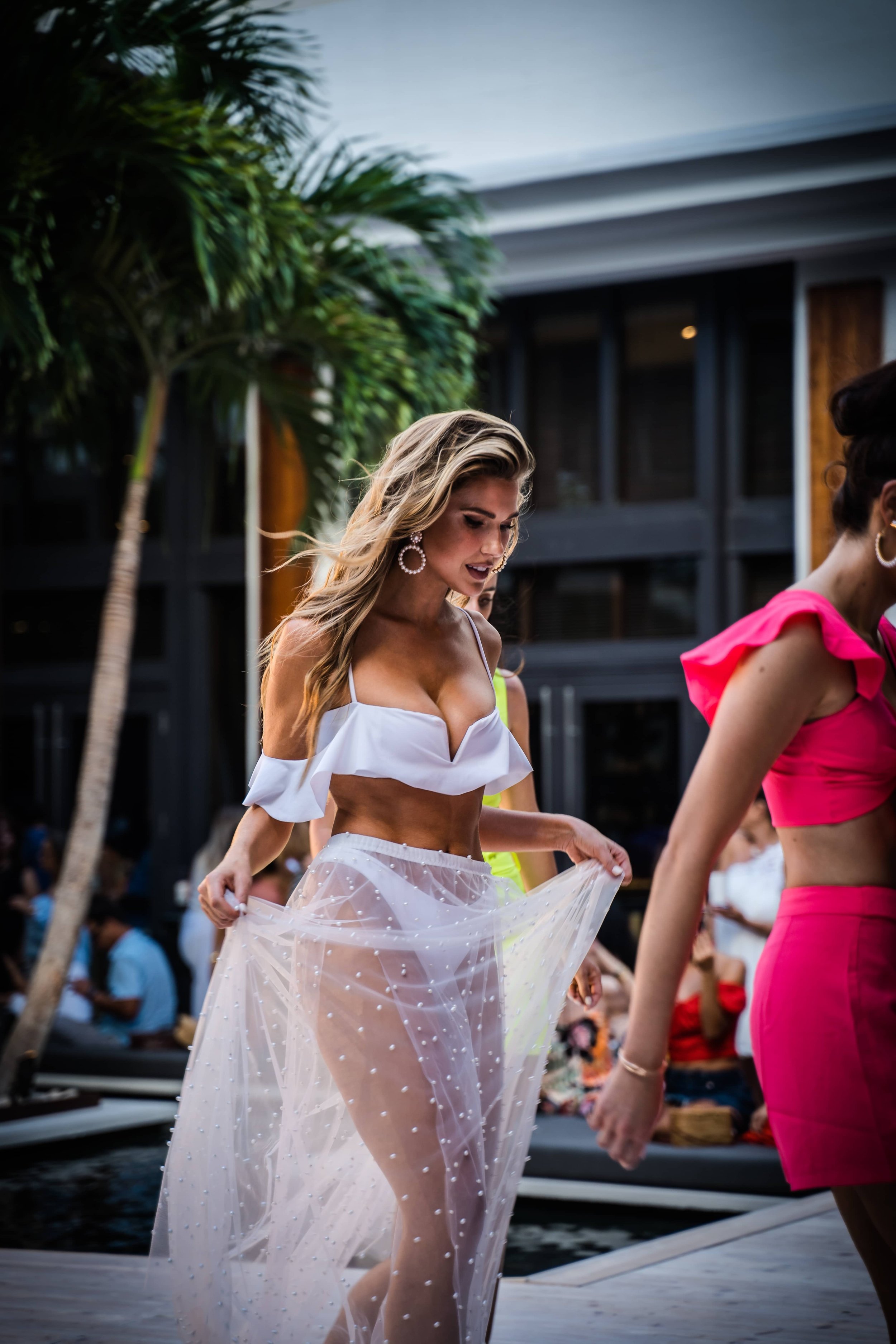 Woman wearing a white crop top and sheer skirt with pearls, holding up her skirt, at a social outdoor gathering with palm trees and people in the background.