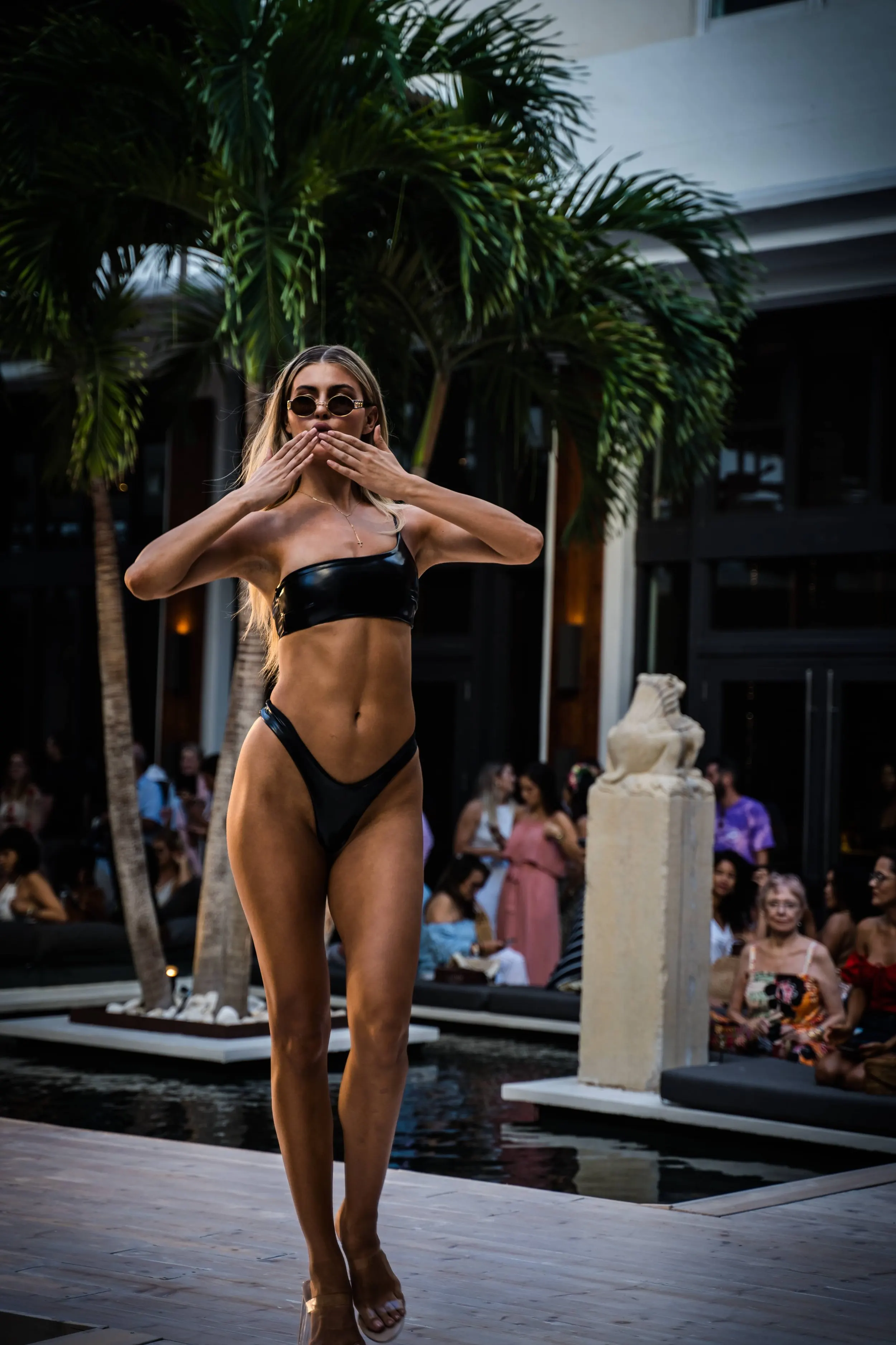 A woman in a black bikini and sunglasses posing at a poolside fashion show, with an audience and palm trees in the background.