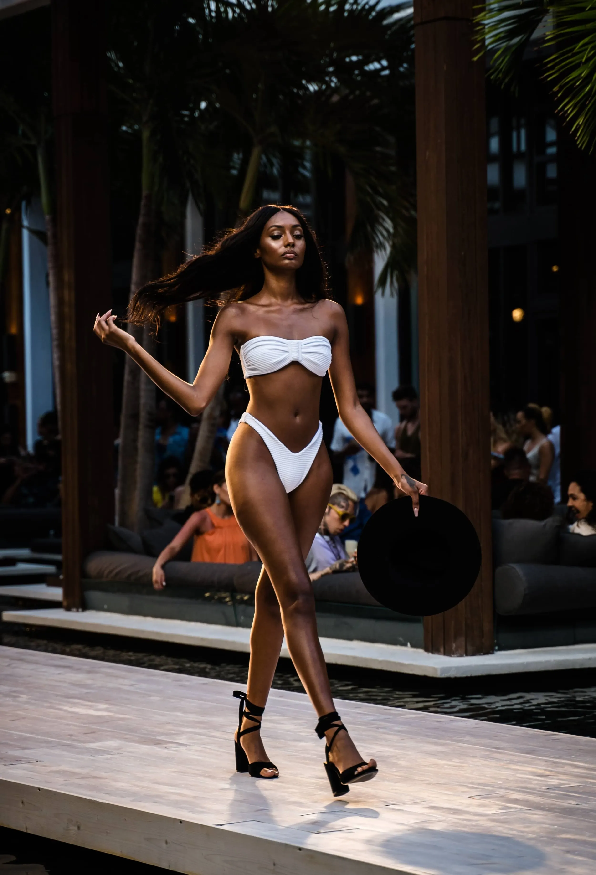 A woman walking on a runway in a white bikini and black high heels, holding a black hat, during a fashion show at an indoor venue with wooden beams and audience in the background.