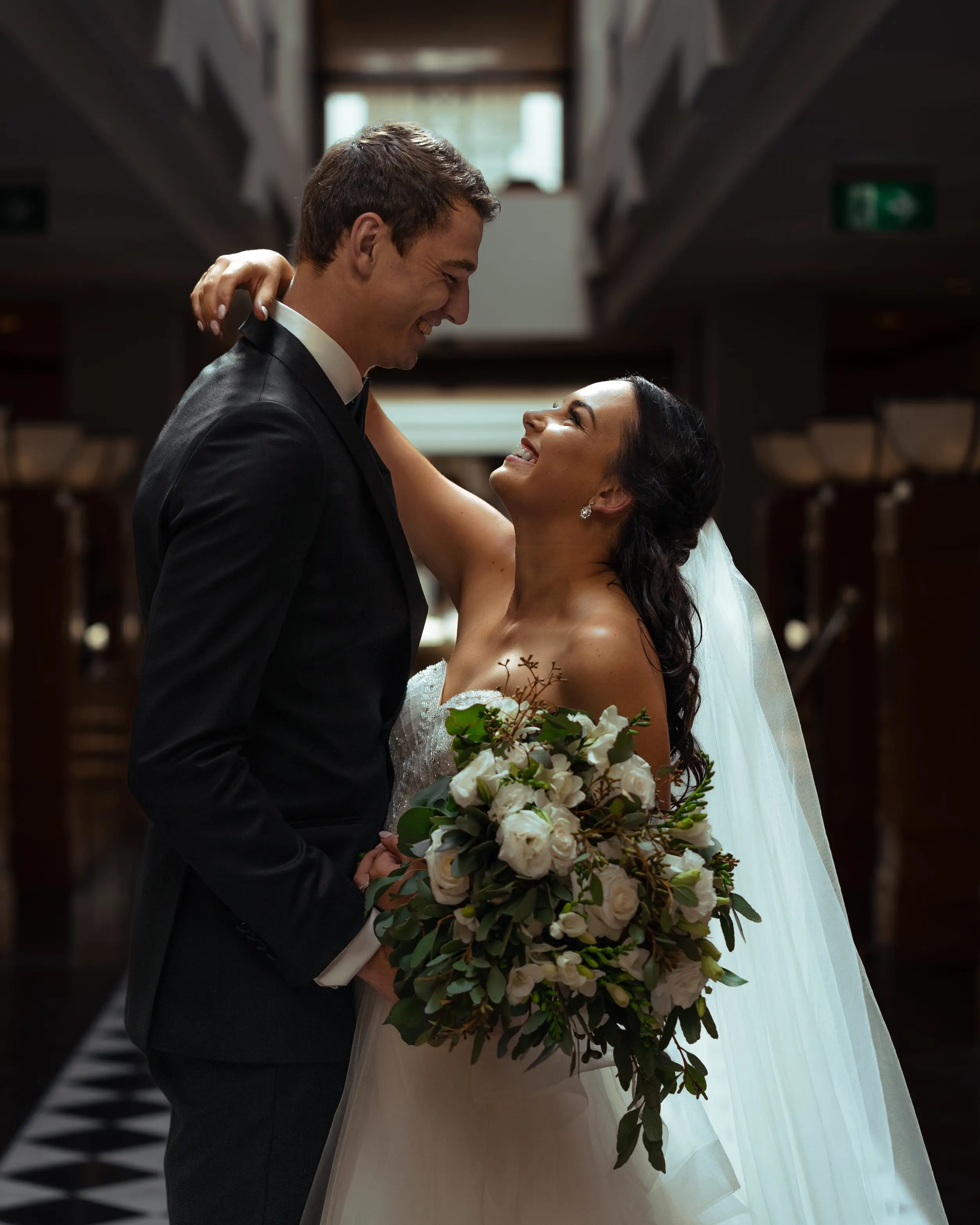 A bride and groom smiling at each other, the bride holding a bouquet of white flowers, in a dimly lit indoor setting.
