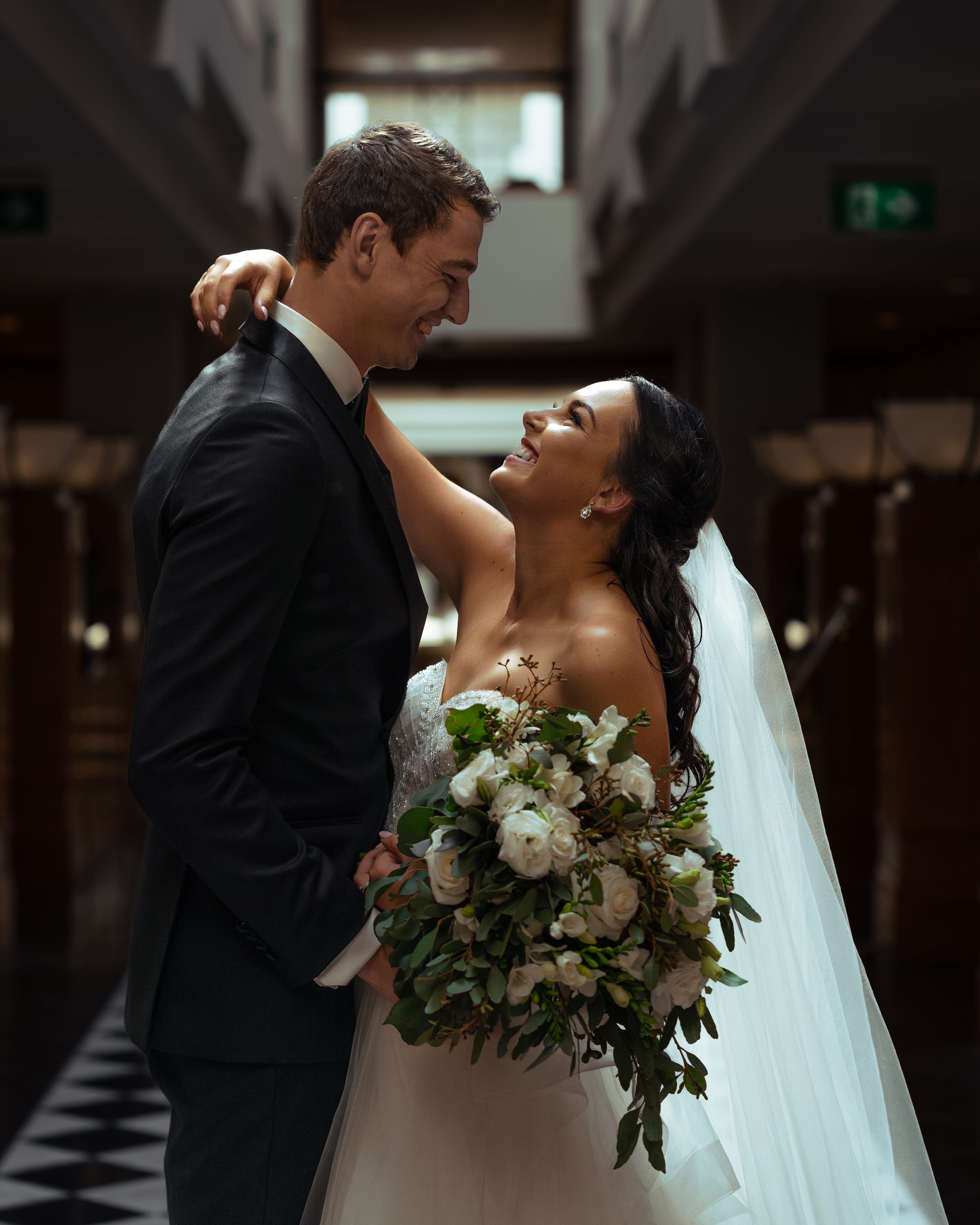 A bride and groom smiling at each other, the bride holding a bouquet of white flowers, in a dimly lit indoor setting.