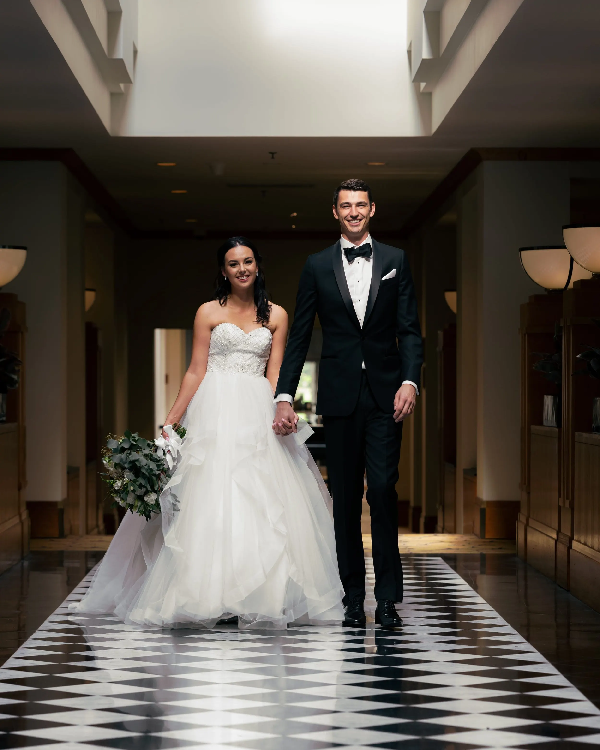 A bride and groom walking hand in hand in an indoor hallway, smiling. The bride is wearing a white strapless wedding gown and holding a bouquet, while the groom is dressed in a black tuxedo with a bow tie.