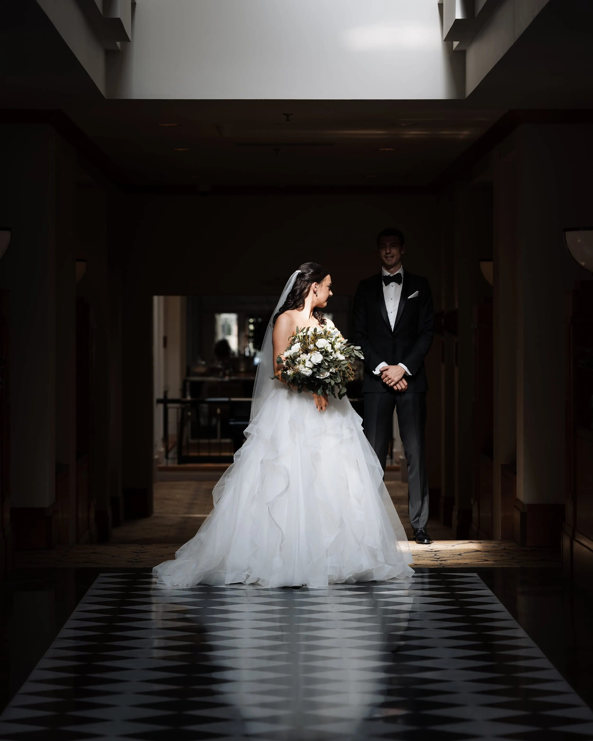 A bride and groom stand in a dimly lit hallway with a skylight above. The bride wears a white wedding gown and holds a bouquet, while the groom wears a black tuxedo and bowtie, smiling at each other.