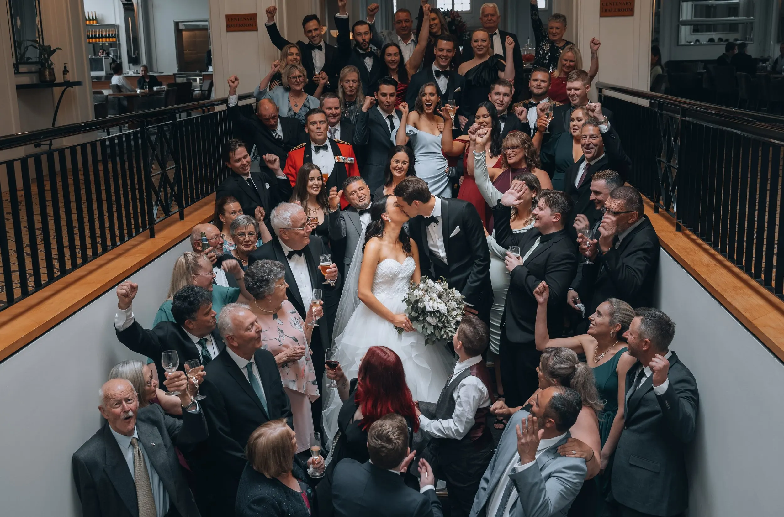 A large group of wedding guests gathered around a bride and groom who are kissing, with many guests raising their glasses and fists in celebration in a hotel lobby or event space.