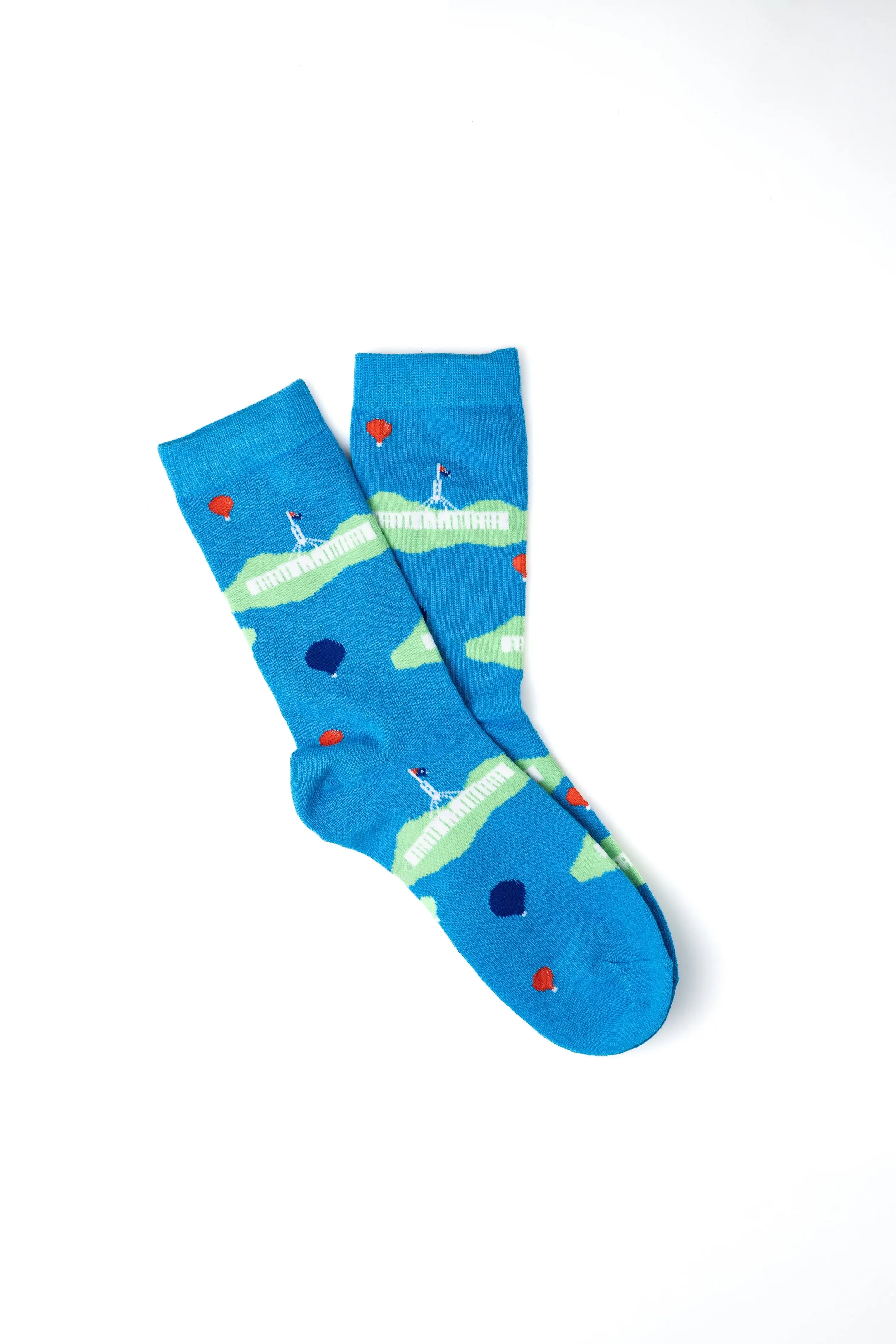 A pair of blue socks featuring a lighthouse, green grass, red and blue rocks, and a small house, placed on a white background.