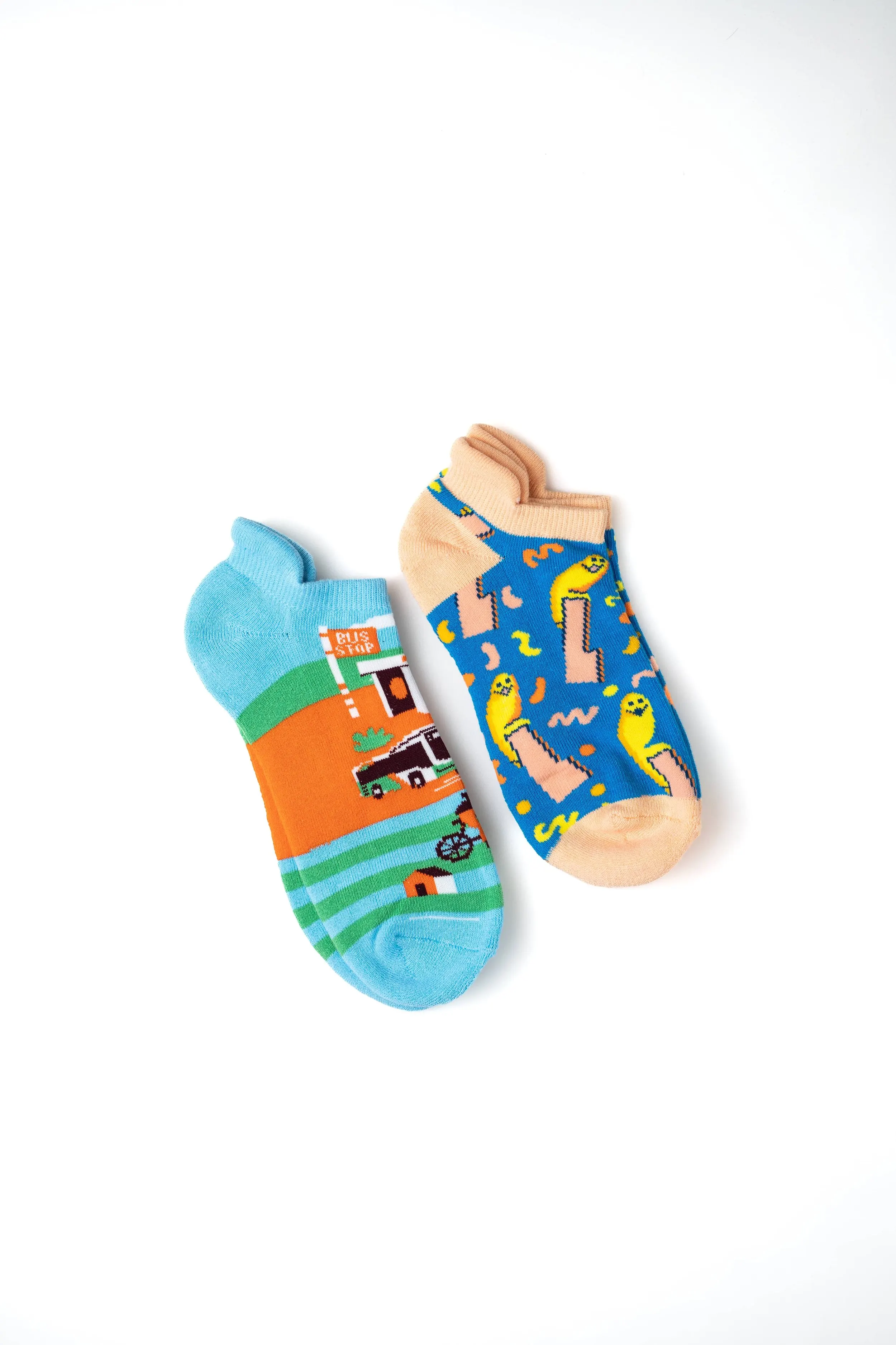 Two colorful children's socks with playful and cartoonish patterns, one featuring a landscape with a bus, house, and trees, and the other with abstract shapes and yellow characters, placed on a white background.