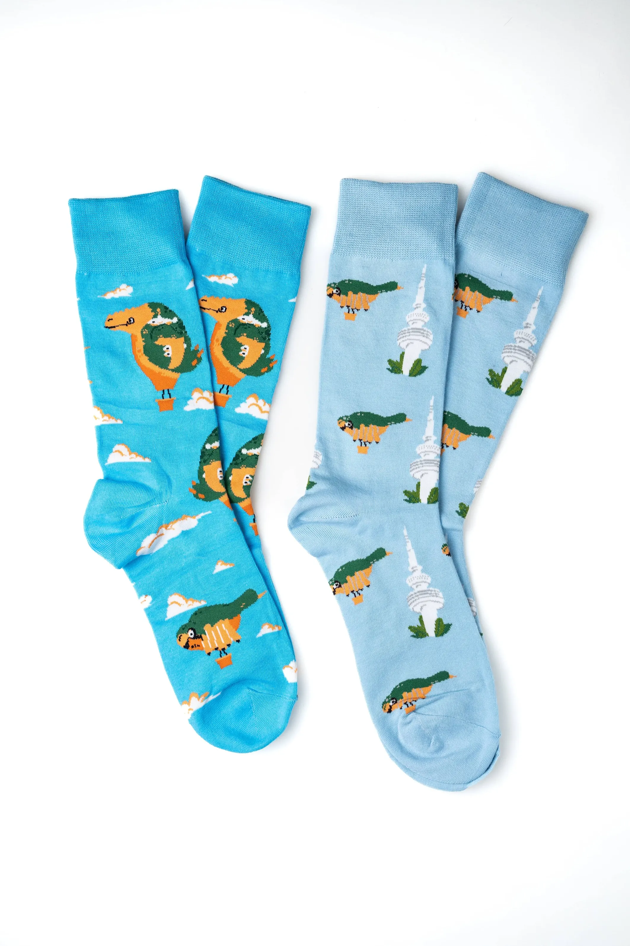 Two pairs of colorful socks with dinosaur designs, one with flying dinosaurs among clouds and the other with dinosaurs standing near a building, on a white background.