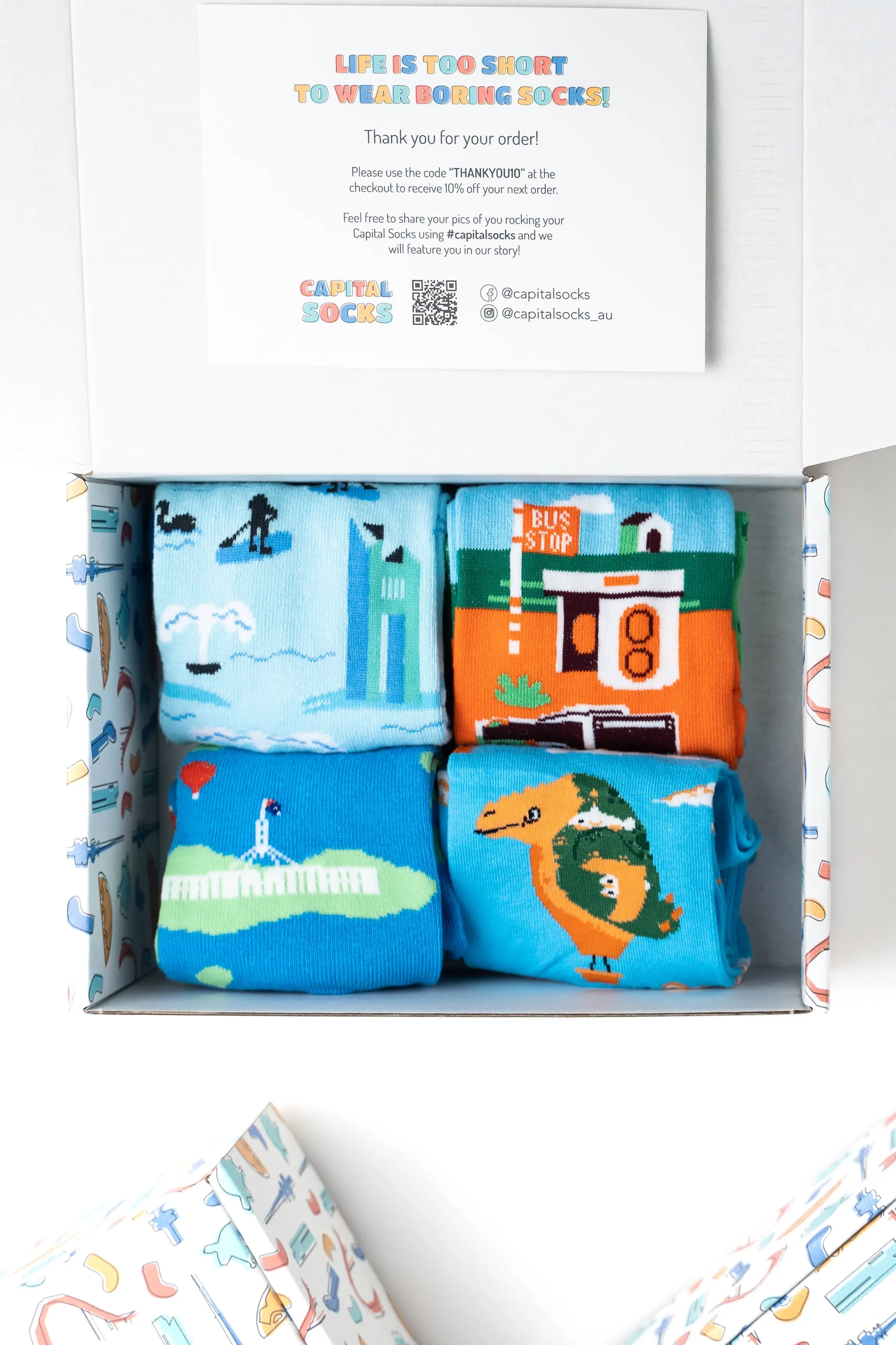 A white box containing four folded colorful socks with pixel art designs, including a cityscape, a bus stop, a lighthouse, and a dinosaur.