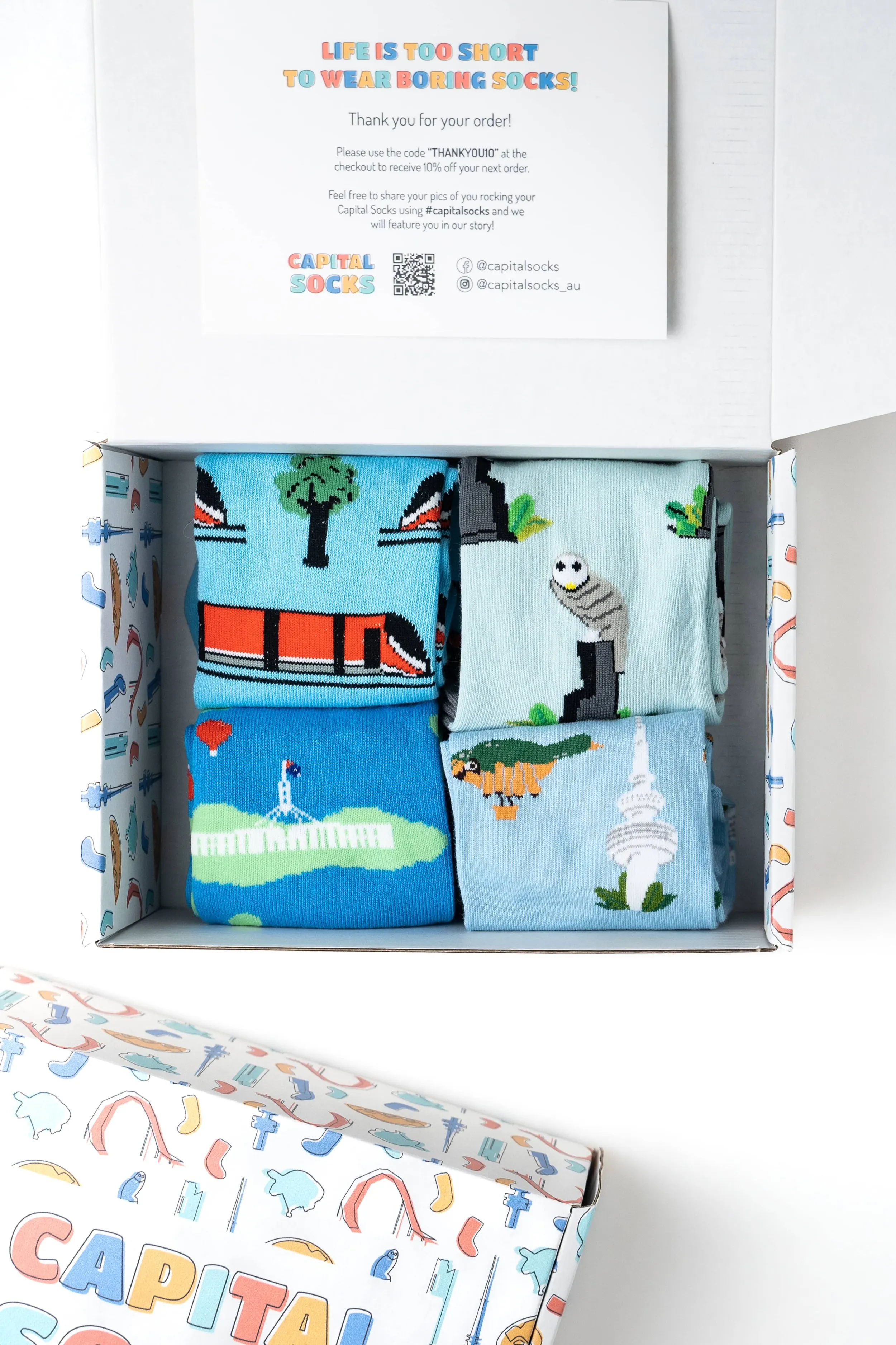 A white box containing four colorful cartoon-style socks with various pixelated landmarks and objects, including a bridge, a lighthouse, a spaceship, and a tower, with a thank you card above the box and the box lid partially open showing more sock pa