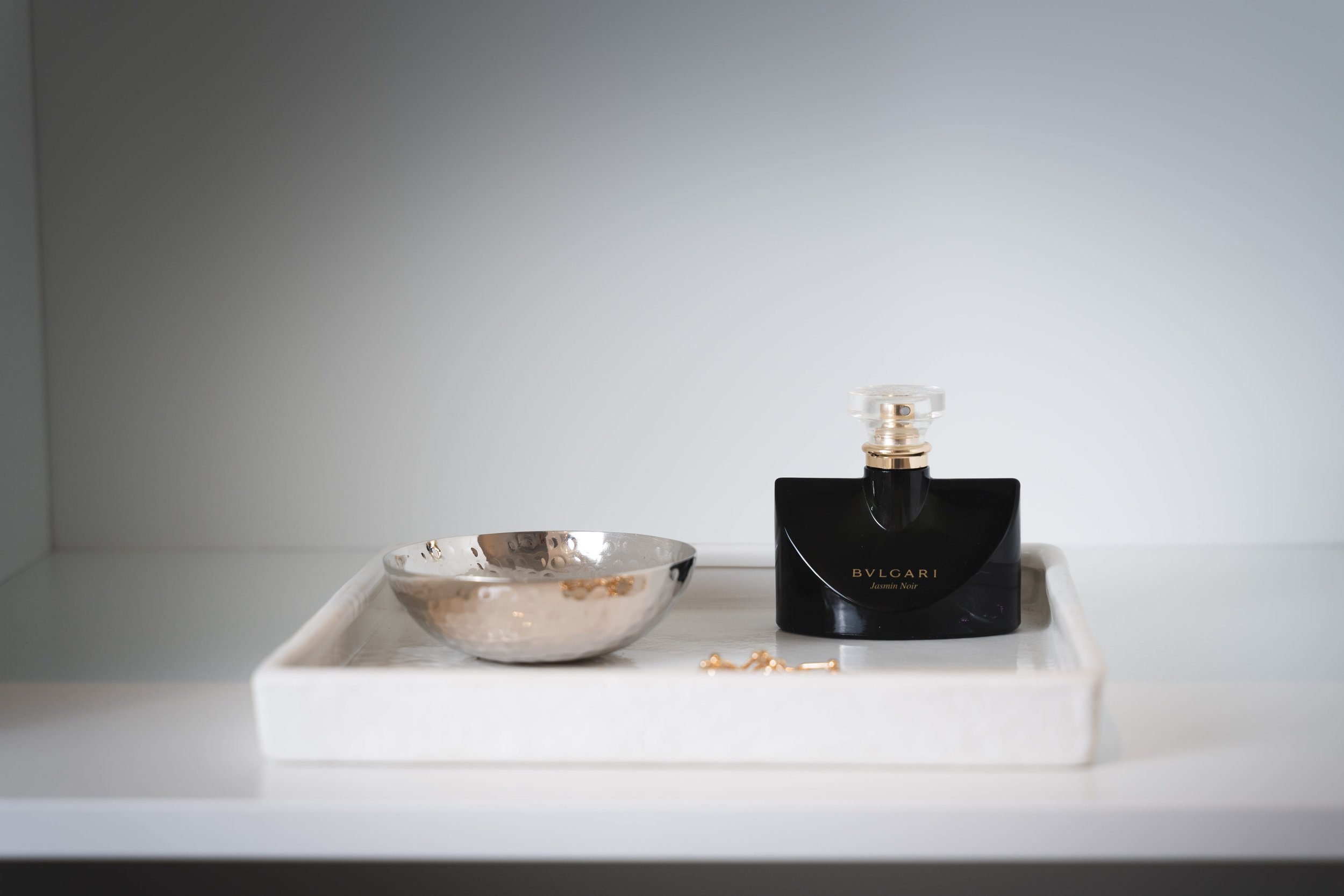 Bvlgari Jasmin Noir perfume bottle on a white tray, with a small metallic bowl and a gold chain bracelet.